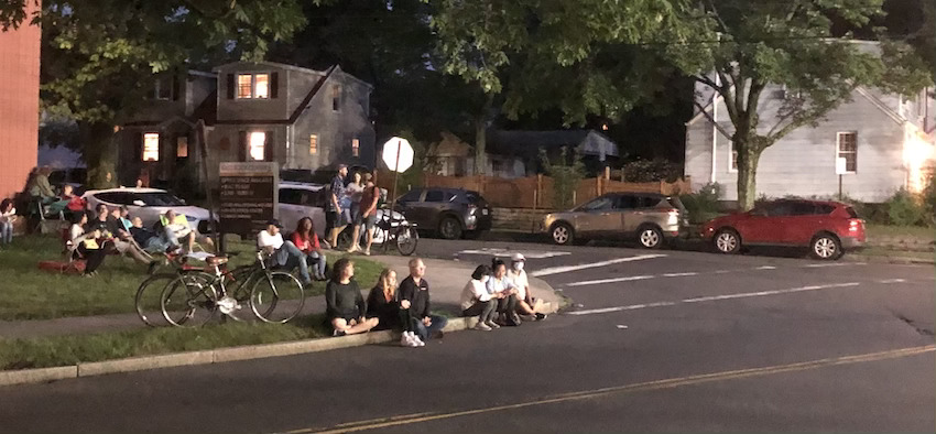 A group of people sitting at a street corner, on the curb, and in chairs.