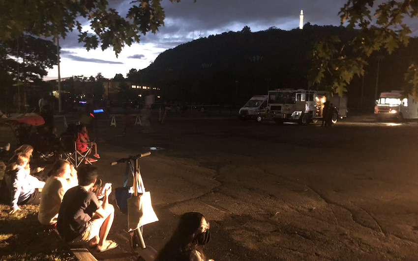 Several people sitting on a parking lot curb, looking at the sky over East Rock Park.
